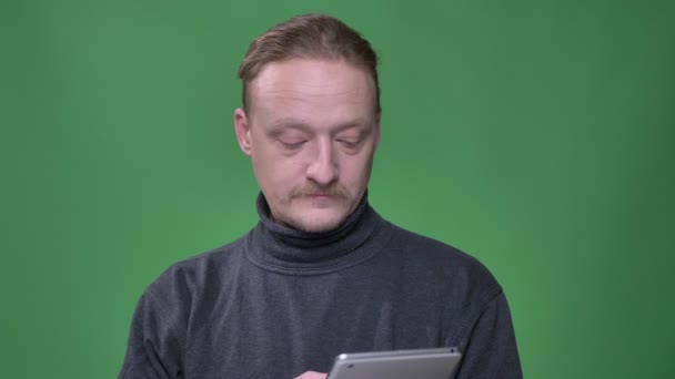 Blonde pensioner in gray pullover works attentively into tablet and demonstrates blue screen of it on green background. — Stock Video