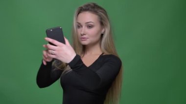 Yeşil arka planda akıllı telefon kullanarak selfie fotoğrafları çeken siyah bluzlu genç modelin portresi.