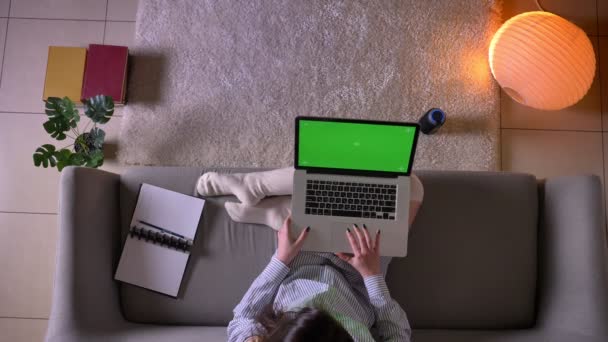 Hlavní stříšku mladé atraktivní ženy používající notebook se zelenou obrazovkou, která sedí na gauči v roztomilém ponožku v útulném bytě — Stock video