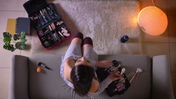 Крупним планом знімок молодої привабливої жінки, що наносить макіяж, сидячи на дивані в милих шкарпетках в приміщенні в затишній квартирі — стокове відео