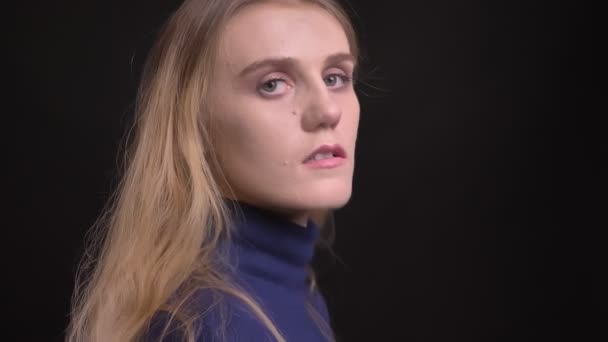 Jong blond model in blauw trui draait naar camera en kijken serieus in het op zwarte achtergrond. — Stockvideo