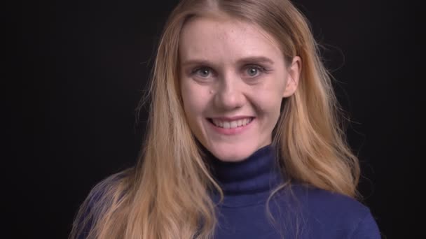 Молодая блондинка в голубом свитере смотрит с искренней и радостной улыбкой в камеру на черном фоне . — стоковое видео