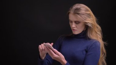 Genç sarışın model akıllı telefondan fotoğraf araklıyor ve siyah arka planda duygusal tepki veriyor..