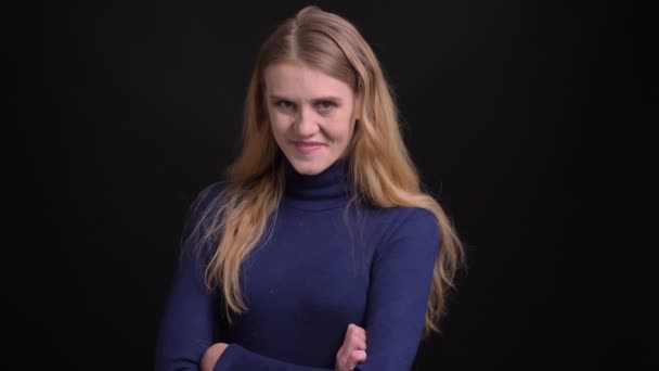 Junges blondes Model im blauen Pullover schaut mit schüchternem und hübschem Lächeln auf schwarzem Hintergrund in die Kamera. — Stockvideo