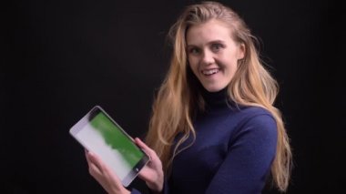 Tabletle çalışan genç sarışın model, siyah arkaplan uygulamasını tavsiye etmek için yeşil ekranını kameraya gösteriyor.