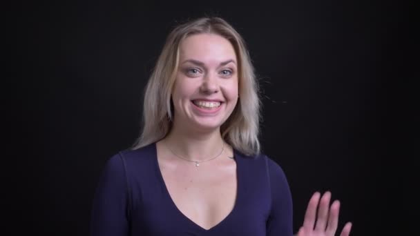 Счастливая блондинка средних лет в синей блузке улыбаясь машет рукой в камеру на черном фоне . — стоковое видео