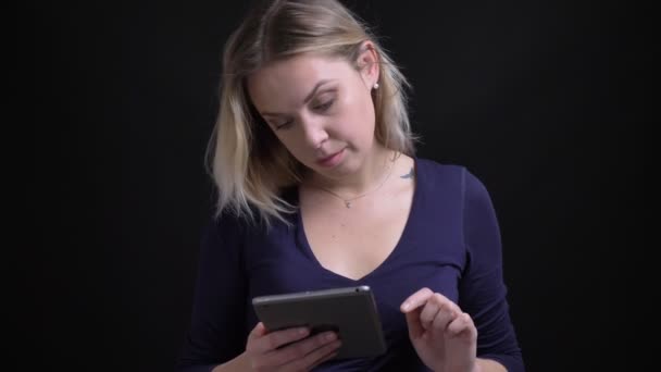 Blonde zakenvrouw in blauwe blouse werken zorgvuldig met Tablet horloges in de camera serieus op zwarte achtergrond. — Stockvideo