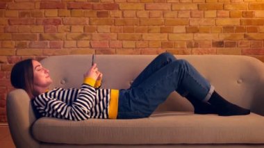 Telefonu kullanarak genç güzel kız Closeup yan görünüm portre ve neşeyle kapalı rahat bir dairede kanepede rahat yatan gülüyor