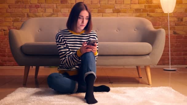 Retrato de cerca de la joven chica bonita usando el teléfono sentado en el suelo en un acogedor apartamento en el interior — Vídeos de Stock