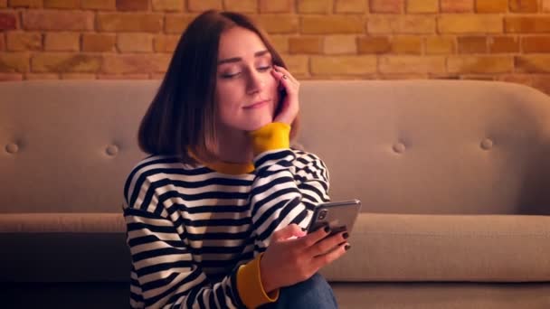 Close-up portret van jonge mooie meisje browsen sociale media op de telefoon en glimlachend gelukkig zittend op de vloer in een gezellig appartement binnenshuis — Stockvideo