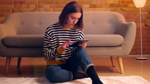 Retrato de cerca de la joven chica bonita usando la tableta sentada en el suelo en un acogedor apartamento en el interior — Vídeo de stock