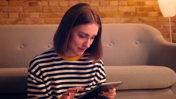 Retrato de close-up da jovem menina bonita digitando no tablet e sorrindo feliz sentado no chão em um apartamento acolhedor dentro de casa — Vídeo de Stock