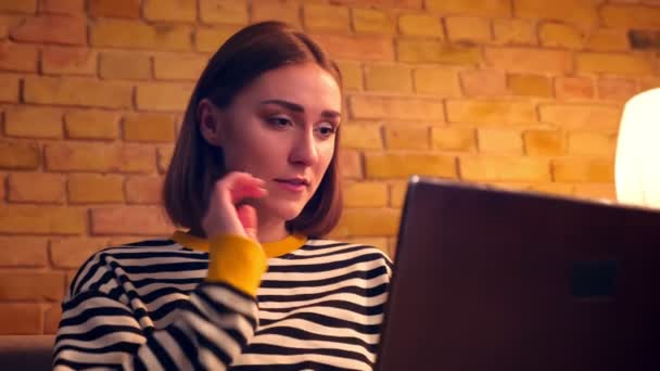 Primer plano retrato de la joven adolescente bonita usando el portátil y riendo alegremente sentado en el sofá en un acogedor apartamento en el interior — Vídeos de Stock