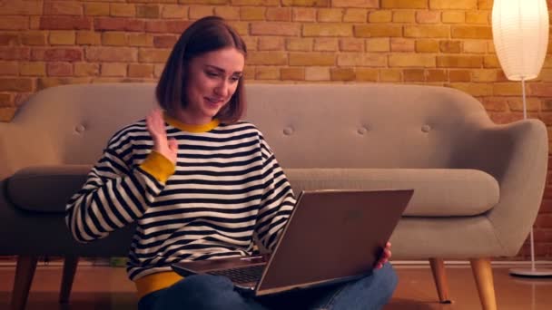 Dizüstü bilgisayarda bir görüntülü arama olan genç güzel kız Closeup portre ve merhaba mutlu kapalı rahat bir dairede katta oturan gülümseyerek sallayarak — Stok video