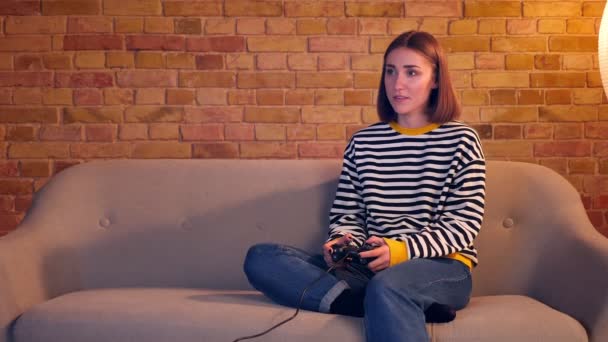 Primer plano retrato de una chica bonita joven jugando videojuegos usando la consola de juegos sentada en el sofá en un acogedor apartamento interior — Vídeos de Stock