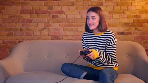 Primer plano retrato de una joven chica bonita jugando videojuegos usando la consola de juegos con emoción sentada en el sofá en un acogedor apartamento interior — Vídeos de Stock