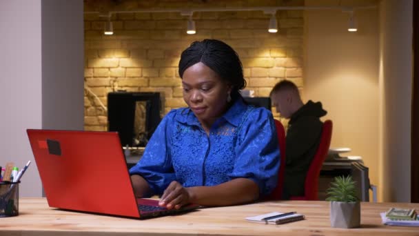 Nahaufnahme einer erwachsenen afrikanisch-amerikanischen Geschäftsfrau, die ihren Laptop benutzt und im Büro am Arbeitsplatz in die Kamera lächelt — Stockvideo