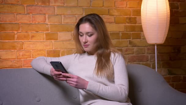 Close-up retrato de jovem atraente caucasiano morena feminino usando o telefone situado no sofá dentro de casa em um apartamento aconchegante — Vídeo de Stock