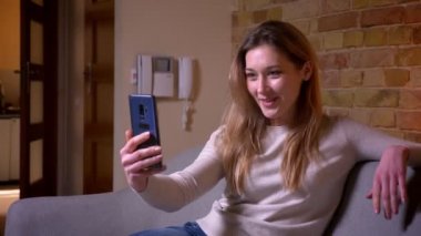 Genç çekici beyaz esmer kadın rahat bir dairede kanepede oturarak kamera önünde poz telefonda selfie çekerken Closeup portre