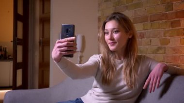 Genç çekici beyaz esmer kadın rahat bir dairede kapalı kanepede oturarak kamera önünde poz cep telefonu selfie çekerken closeup portre