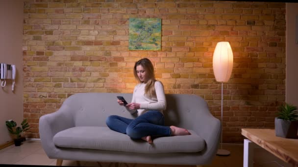 Крупним планом портрет молодої привабливої білоруської брюнетки жіночий перегляд по телефону, сидячи на дивані в приміщенні в затишній квартирі — стокове відео