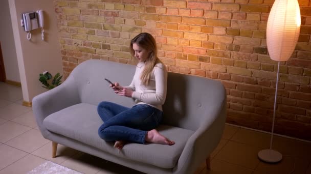 Closeup portrait of young attractive caucasian brunette female texting the phone siting on the sofa indoors in a cozy apartment — Stock Video