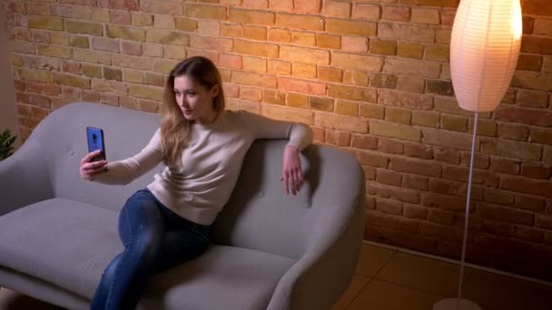 Close-up retrato de jovem atraente caucasiano morena fêmea tomando selfies no telefone sorrindo feliz situando no sofá dentro de casa em um apartamento aconchegante — Vídeo de Stock