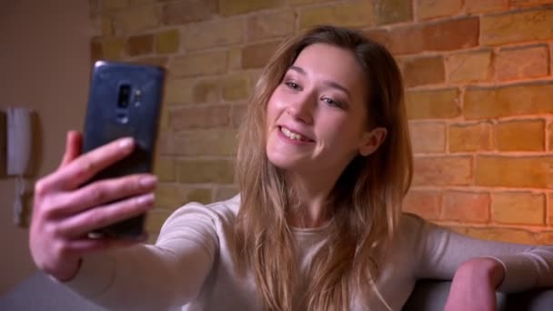 Closeup portrait of young attractive caucasian brunette female taking selfies on the cellphone posing in front of the camera siting on the sofa smiling happily — Stock Video