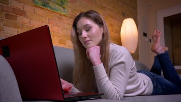 Närbild porträtt av unga attraktiva kaukasiska brunett affärskvinna arbetar fjärrkontroll på den bärbara datorn och liggande på soffan inomhus i en mysig lägenhet — Stockvideo