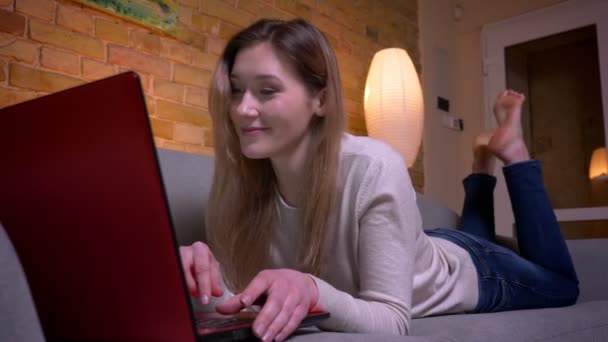 Крупним планом портрет молодої привабливої кавказької брюнетки жіночих повідомлень на ноутбуці і прослуховування музики з радістю лежить на дивані в приміщенні в затишній квартирі — стокове відео