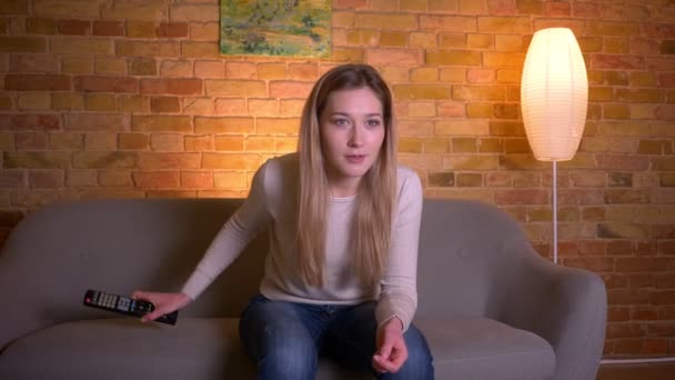 Nahaufnahme Porträt der jungen attraktiven kaukasischen brünetten Frau vor dem Fernseher und glücklich Sitzen auf der Couch drinnen in einer gemütlichen Wohnung — Stockvideo