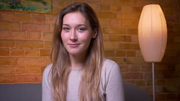 Close-up portret van jonge schattige Kaukasische brunette vrouw kijken naar camera en glimlachend gelukkig plaatsings op de Bank binnenshuis in een gezellig appartement — Stockvideo