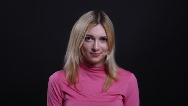 Close-up shoot van jonge mooie blonde vrouw glimlachend met verlegenheid en verwarring kijkend naar camera met achtergrond geïsoleerd op zwart — Stockvideo
