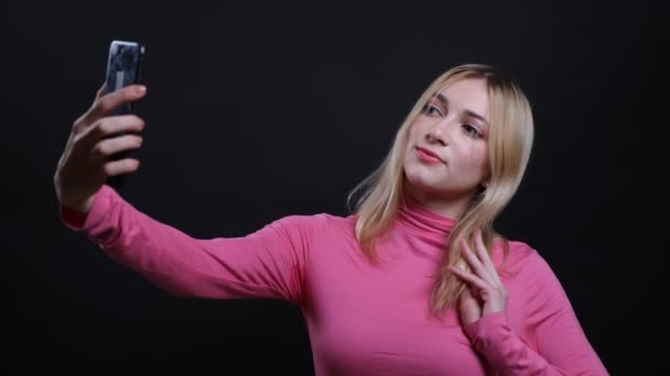 Close-up shoot van jonge mooie blonde vrouw die selfies neemt op de telefoon en glimlacht gelukkig met achtergrond geïsoleerd op zwart — Stockvideo