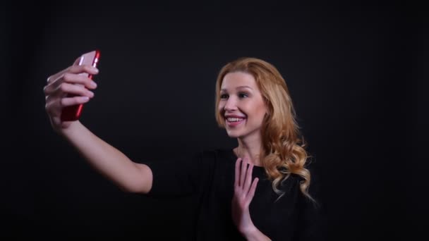 Крупный план портрет взрослой привлекательной рыжеволосой женщины с видеозвонком по телефону, машущим привет и весело говорящим на фоне, изолированном от черного — стоковое видео