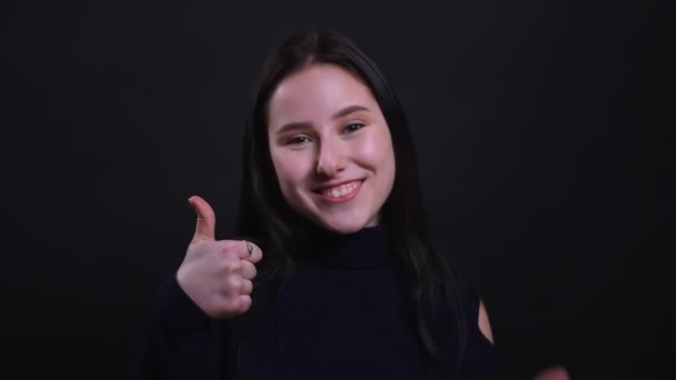Close-up retrato de jovem atraente morena feminino mostrando um polegar para cima sorrindo com confiança olhando para a câmera com fundo isolado no preto — Vídeo de Stock