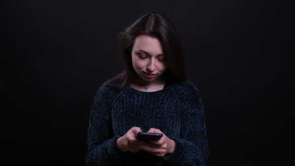 Портрет крупным планом взрослой красивой брюнетки кавказской женщины с телефоном и смотрящей в камеру на фоне изолированного на черном фоне — стоковое видео