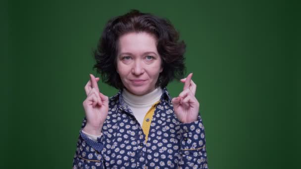 Крупный план портрет старой брюнетки белого цвета женщины со скрещенными пальцами беспокоясь и глядя в камеру с изолированным на зеленом фоне — стоковое видео