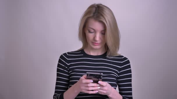 Primer plano retrato de adulto atractivo rubia caucásica femenina mensajes de texto en el teléfono y sonriendo con fondo aislado en blanco — Vídeos de Stock