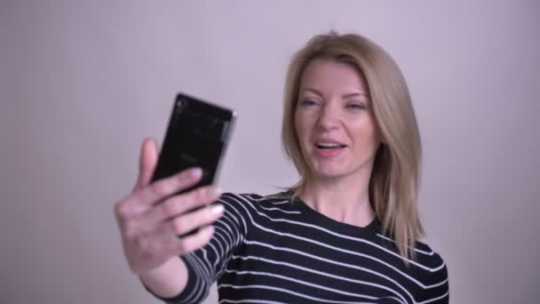 Closeup portrét dospělé atraktivní blond kavkazské samice, která má video hovor na telefonu a usmívá se na pozadí izolovaně na bílém — Stock video