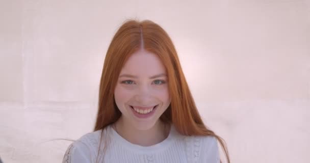 Портрет симпатичной имбирной модели поворачивается к камере и радостно улыбается ей на фоне белой стены . — стоковое видео