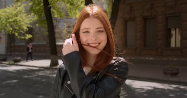 Deri ceketli genç kızıl kız kameraya güzelce gülümsüyor ve saçını şehrin arka planında düzeltiyor..