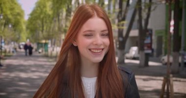 Deri ceket güzel zencefil kız kameraya döner ve yeşil şehir sokak arka planda kameraya mutlu gülümsüyor.