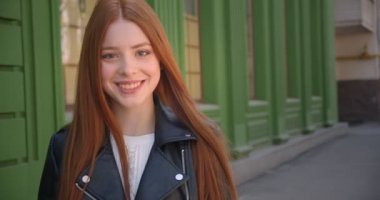 Deri ceket güzel zencefil kız Yürüyüş yeşil ev arka planda içine gülümsüyor kamera önünde durur.