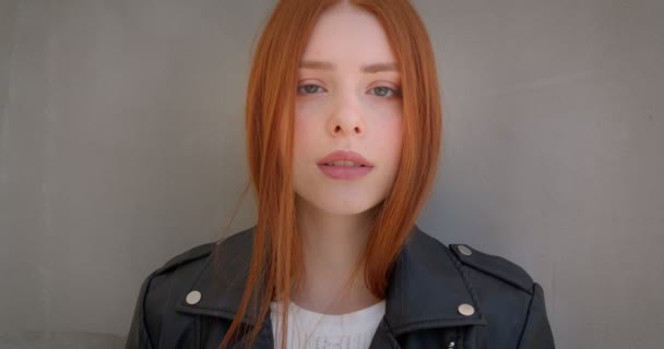 Young female ginger rocker in leather jacket flirting with camera leaning on the white wall. — ストック動画