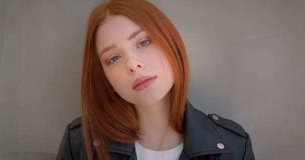 Young female ginger rocker in leather jacket watching calmly into camera leaning on the white wall. — ストック動画