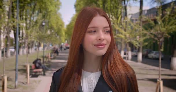 Belle fille roux en veste en cuir regardant joliment dans la caméra sur fond de ruelle verte de la ville . — Video