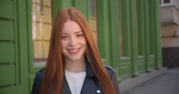 Camminando bella ragazza con la testa rossa in giacca di pelle si ferma di fronte alla fotocamera sorride in esso su sfondo verde casa . — Video Stock
