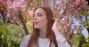 Pembe çiçekli bahçede kameraya rüya gibi izlerken beyaz bluz büyüleyici zencefil kız öğrenci.