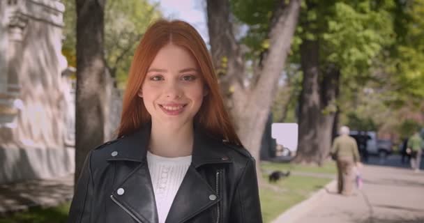 Caminhando menina gengibre bonita em jaqueta de couro pára na frente da câmera sorri para ele no fundo do parque verde . — Vídeo de Stock